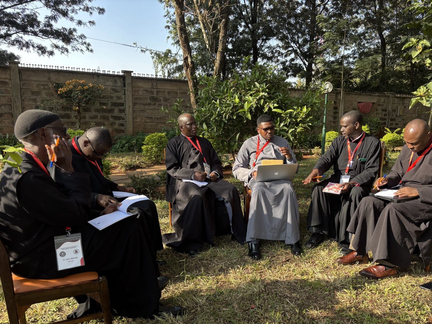A writing group sitting in a circle of chairs out on the grass, papers and a laptop on their knees.