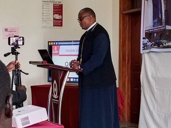 Fr Thiani presenting a paper at a lectern in a university lecture hall.