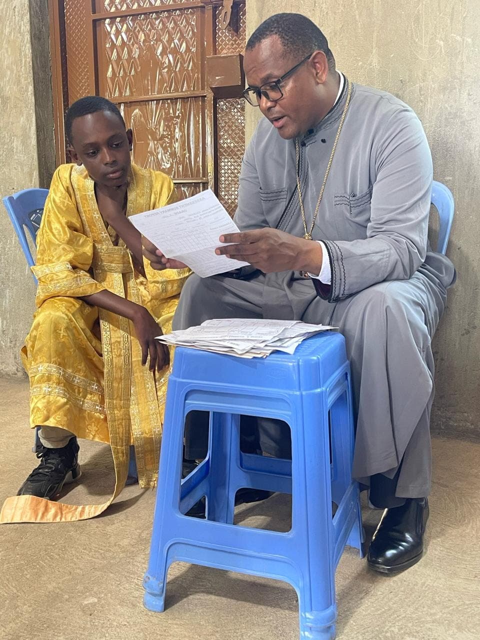 Fr Thiani reading through a school report with an altar server in gold vestments, a stack of reports on the stool between them.