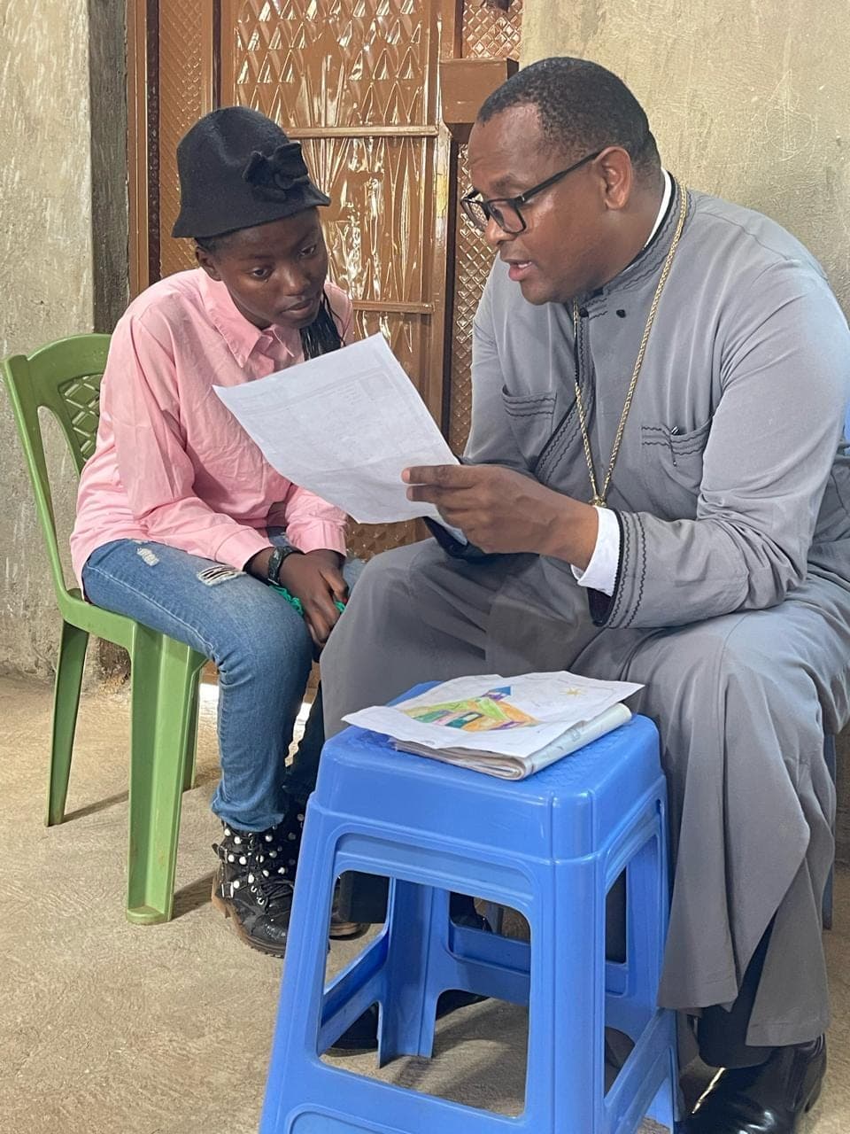 Fr Thiani talking a student through her marks, her exercise book open on the stool beside them.