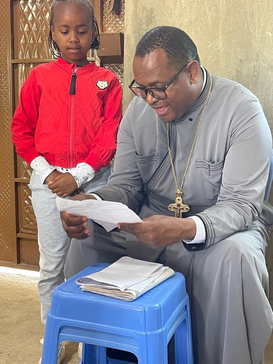 A young girl in a red jacket standing beside Fr Thiani as he reads out her report.